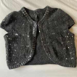 Grå paljettväst från Cubus - Säljer en mörkgrå stickad väst från Cubus med glittriga paljetter över hela framsidan och längs kanterna. Västen har korta ärmar och rund hals, perfekt för att ge din outfit extra edge. Materialet är mjukt och fluffigt, vilket gör den superbekväm.