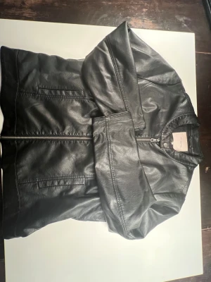 Svart skinnjacka från Only Carmakoma - Snygg svart skinnjacka från Only Carmakoma med klassisk rund krage och dragkedja framtill. Jackan har diskreta fickor på sidorna och en metallknapp vid halsen med logga. Perfekt för dig som vill ha en cool och tidlös look.