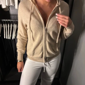 Beige hoodie i kashmir från Soft Goat - Mjuk och lyxig beige hoodie från Soft Goat i kashmir. Tröjan har dragkedja framtill, huva med snörning och ribbade muddar. Perfekt nu till hösten och vintern. Passformen är normal och materialet är supermjukt. Har en liten fläck vid ärmen som går bort i kemkvätt (därav priset 