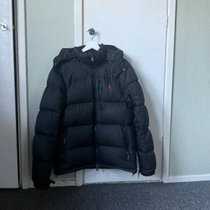 Svart pufferjacka från Polo Ralph Lauren - Svart pufferjacka från Polo Ralph Lauren med huva och flera dragkedjeförsedda fickor. Jackan har det klassiska röda Polo-logot broderat på bröstet och är fodrad för extra värme. Perfekt för kalla dagar och har en clean, sportig look.