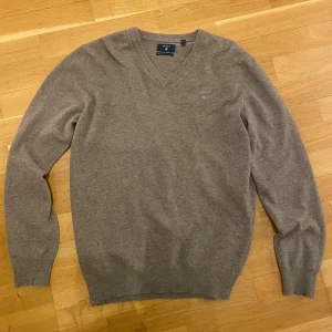 Beige v-ringad tröja från Gant - Snygg beige v-ringad tröja från Gant i mjuk ullmix i storlek L men kan användas som M. Klassisk design med ribbade muddar vid ärmslut och nederkant samt diskret logga broderad på bröstet. Perfekt för dig som gillar stilrena och tidlösa plagg.