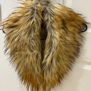 Fuskpäls halsduk från Saki - Supermjuk och fluffig fuskpäls halsduk från Saki i snygga toner av beige, brun och svart. Halsduken har långhårig pälslook och är tillverkad i polyester. Perfekt för att ge din outfit en extra edge under kyliga dagar.