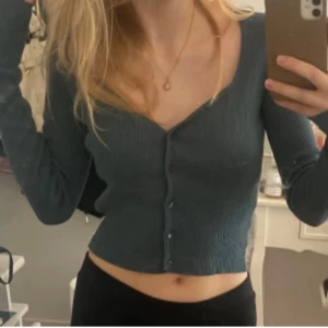 Grön ribbad croppad topp från H&M - Säljer en grön ribbad croppad topp från H&M i storlek XS. Toppen har lång ärm, v-ringad hals och knappar framtill. Snygg slim passform som framhäver figuren och passar perfekt till jeans eller kjol. Materialet är mjukt och stretchigt för en bekväm känsla.