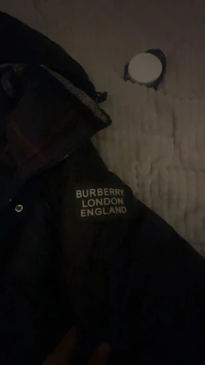 Svart pufferjacka från Burberry - Svart pufferjacka från Burberry med huva och klassiskt rutigt foder i kragen. Jackan har dragkedja och knappar framtill, samt två fickor med dragkedja. På vänster ärm finns Burberry London England-tryck. Perfekt för kalla dagar.