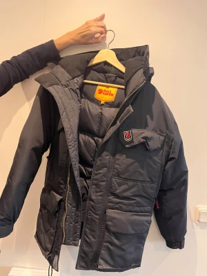 Svart dunjacka från Fjällräven XXXS - Svart dunjacka från Fjällräven med klassisk röd logga på bröstet och gul insida vid etiketten. Jackan har huva, flera fickor med lock och dragkedja framtill. Perfekt för kalla dagar och har en schysst, puffig look som är både praktisk och snygg.  XXXS herr storlek motsvarar storleken S