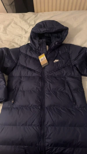 Mörkblå Nike pufferjacka med huva - Säljer en mörkblå pufferjacka från Nike med huva och två sidofickor med dragkedja. Jackan har en stilren design med Nike-logga på bröstet och quiltade paneler. Perfekt för kalla dagar och enkel att matcha med din streetwear. Och har använt den 1 