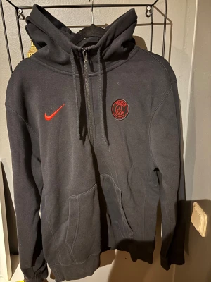 Svart PSG hoodie från Nike med röd print - Svart hoodie från Nike med Paris Saint-Germain-logga. Stor röd print med siffror och fleur-de-lis på ryggen. Tröjan har huva, dragkedja och två fickor framtill. 