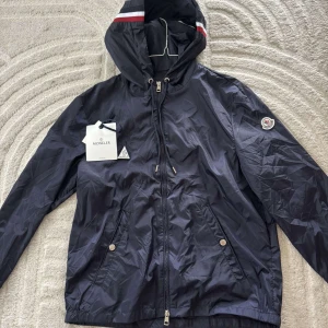 Svart vindjacka från Moncler med huva - Snygg svart vindjacka från Moncler med huva och dragkedja framtill. Jackan har Moncler-logga på ärmen, två fickor med knappar och coola ränder på huvan. Perfekt för dig som vill ha en stilren och sportig look. Men ett litet hål i ryggen som nt man ser ifrån långt håll men annars jätte fin jacka 