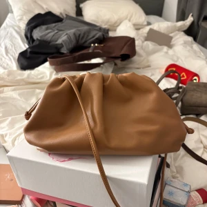 Brun axelväska med snörning - Snygg brun axelväska i mjukt skinn med cool snörning upptill. Väskan har en slouchy form och ett rymligt fack, perfekt för dina essentials. Stilren design med långa axelband och knutdetaljer på sidorna. Passar dig som gillar minimalistisk och trendig look.