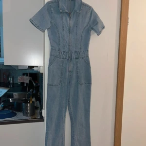 Ljusblå jeans jumpsuit med dragkedja  - Snygg ljusblå jumpsuit i denim med korta ärmar och markerad midja. Framtill finns en dragkedja hela vägen upp till kragen och två stora fickor. Perfekt för dig som gillar en cool och avslappnad stil.