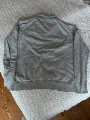 Grå half zip tröja från Gant - Snygg grå långärmad tröja från Gant med half zip och broderad logga på bröstet. Tröjan har ribbade muddar och krage med diskreta svarta ränder. Perfekt för dig som gillar stilren och klassisk look.