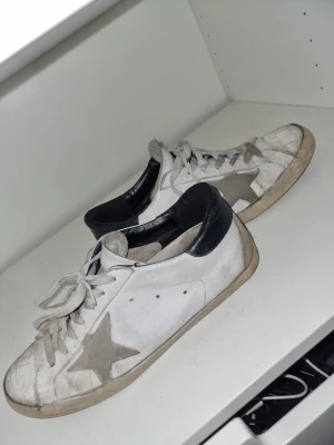 Golden Goose vita sneakers med stjärna - Golden Goose sneakers i vitt skinn med grå mockadetaljer och en stor grå stjärna på sidan. Svart hälparti och platt sula. Klassisk låg modell med snörning och rund tå. Perfekt för dig som gillar en cool och avslappnad stil. Ett G på sidan där det står GGDB ☆ har ramlat av. Först till kvar!