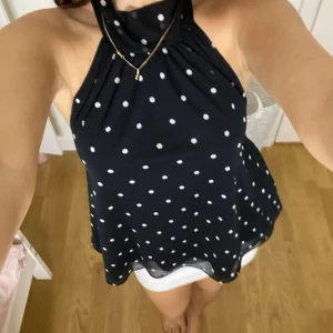 Svart prickig ärmlös linnetopp - Säljer en svart ärmlös linnetopp med vita prickar och hög hals. Toppen har en flowy passform och är tillverkad i ett lätt, syntetiskt material som ger en luftig känsla. Perfekt för dig som gillar en stilren men lekfull look.