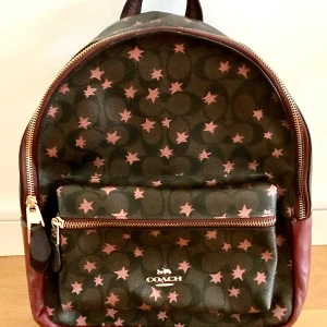 Coach ryggsäck med stjärnmönster - Snygg ryggsäck från Coach i mörkbrunt och vinrött med rosa stjärnor över hela väskan. Väskan har guldfärgade dragkedjor, ett stort huvudfack och en praktisk ytterficka. Materialet är skinn och polyester, med vadderade axelremmar för extra komfort.