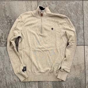 Morris half zip - Snygg beige stickad tröja från Morris med half zip. Tröjan har ett svart broderat emblem på bröstet och ribbade muddar vid ärmslut och nederkant. Nypris är 1600kr. Priset är ej hugget o sten. Hör av dig vid funderingar!