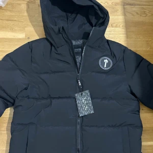 Svart pufferjacka från Trapstar - Svart pufferjacka från Trapstar med huva och dragkedja. Jackan har Trapstar-logga på bröstet och stor broderad text på ryggen. Klassisk pufferdesign med horisontella sömmar och en clean streetwear-look. Perfekt för kalla dagar.