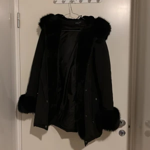 Svart parkajacka med fuskpäls från Hollies - Snygg svart parkajacka från Hollies med stora fuskpälsdetaljer på huva och ärmslut. Jackan har snörning i midjan och stängs med knappar. Perfekt för kalla dagar när du vill vara både varm och stilren.