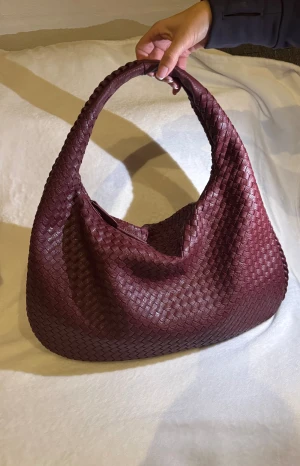 Vinröd flätad axelväska - Snygg vinröd axelväska med flätad design och mjuk, rundad form. Design by Si- Musthave bag i vinröd. Material: vegan leather. Sparsamt använd.