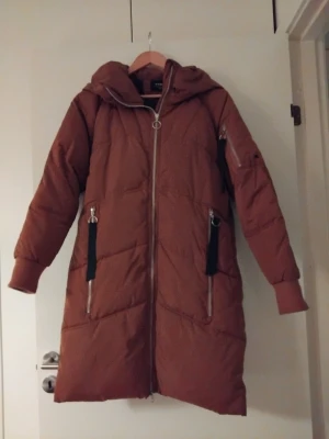 Rostbrun lång pufferjacka med huva - Säljer en rostbrun lång pufferjacka från Everest med huva och snygga silverfärgade dragkedjor. Jackan har svarta detaljer vid fickorna och är quiltad för extra värme. Perfekt för kalla dagar och har en modern, loose passform.