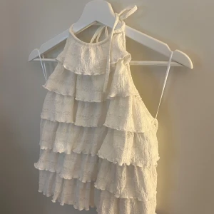 Vit blus med volanger - Supersöt vit blus med flera lager volanger och knyt i nacken. Blusen är ärmlös och har en lätt strukturerad bomullskänsla som ger en chill och luftig vibe. Perfekt för sommarens varma dagar och har en riktigt fräsch look.