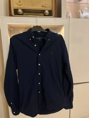 Mörkblå skjorta från Ralph Lauren - Snygg mörkblå skjorta från Ralph Lauren i slim fit-modell. Skjortan har klassisk krage, vita knappar och det ikoniska röda Polo-logot på bröstet. Tillverkad i mjuk bomull som känns skön mot huden. Perfekt för dig som gillar stilrena och tidlösa plagg.