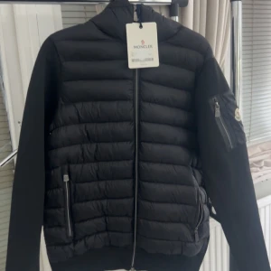 Svart pufferjacka från Moncler - Svart pufferjacka från Moncler med quiltad framsida och huva. Jackan har dragkedja framtill, två sidofickor med dragkedja och en extra ficka med Moncler-logga på ärmen. Materialet är polyester och dun, med en stilren och modern look.