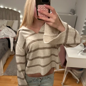 Beige och brun randig stickad tröja - Supermysig stickad tröja i beige med bruna ränder. Tröjan har v-ringning, breda ärmar och croppad passform. Perfekt för lager-på-lager och chill dagar. Snygg till både jeans och kjol.