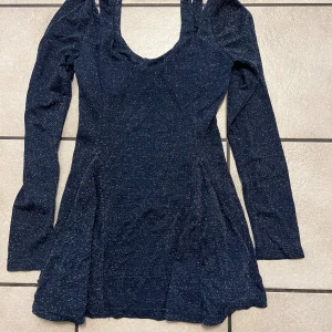 Marinblå glittrig kortklänning storlek XXS/XS - Supersnygg marinblå kortklänning med glittrigt tyg och långärmad design. Klänningen har en rundad urringning och sitter snyggt med en lätt utsvängd kjol. Perfekt för dig som vill sticka ut med lite extra glitter och glam på nästa event.