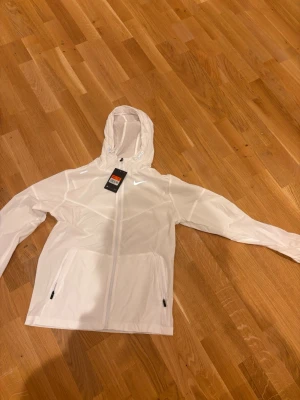 Vit Nike vindjacka med huva, strl M - Snygg vit vindjacka från Nike med huva och dragkedja framtill. Jackan har två fickor med dragkedja och justerbar snörning vid huvan. Perfekt för aktiva dagar och sportiga outfits. Lätt och smidig design med diskret Nike-logga på bröstet.passar folk med S också😊