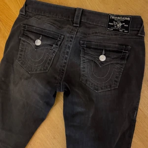 True Religion svarta bootcut jeans - Säljer ett par svarta jeans från True Religion med bootcut passform och washed look. Modellen heter Joey Low Rise Flare och har ikoniska fickor med lock och silverfärgade knappar bak. Jeansen är tillverkade i stretchigt denim.