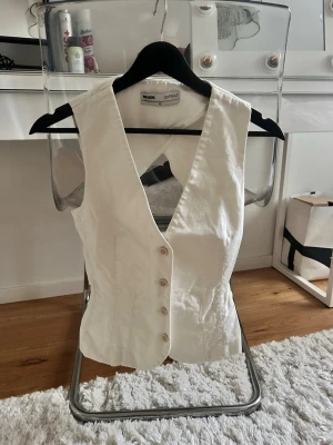 Festtopp från bikbok - Säljer en stilren vit väst från Bik Bok Capsule i storlek XS. Västen har djup v-ringning, knappar framtill och ett bälte bak för extra passform. Materialet känns som bomull och har en klassisk, figurnära siluett som är lätt att matcha. Köptes för 899 och finns inte att köpa längre.