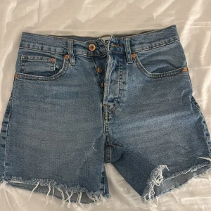 Blå jeansshorts från lager 157 - Säljer ett par blå jeansshorts från lager 157 i storlek S. De har hög midja, klassisk femficksdesign och knappar framtill. Rå kant med fransar nedtill ger en chill vibe. Perfekta för varma dagar och har en loose passform.