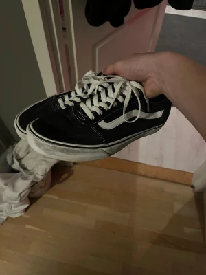 Svarta Vans - Säljer ett par klassiska Vans Old Skool sneakers i svart med vit sula och vit sidostrip. Skorna har snörning och rund tå, med en platt gummisula i brunt. Materialet är en mix av canvas och mocka, vilket ger en schysst streetstil.