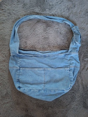 Ljusblå axelväska i denim - Säljer en ljusblå axelväska i denim med bred axelrem och praktisk dragkedja upptill. Väskan har flera fickor och synliga sömmar som ger en cool jeanslook. Perfekt för dig som gillar avslappnad stil och vill ha plats för allt du behöver.