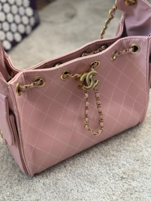 Rosa quiltad axelväska från Ch@nel  - Säljer en rosa axelväska från Ch@nel med klassiskt quiltat mönster och guldfärgade kedjedetaljer. Väskan har CC-logga framtill, sidofickor med lock och snygga metalldetaljer. Insidan har dragkedjefack och väskan är tillverkad i skinn. Perfekt för dig som gillar lyxiga accessoarer.