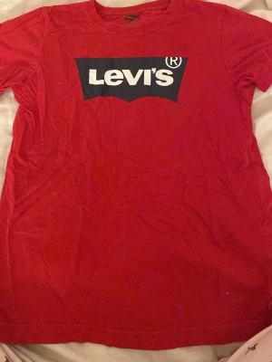 Röd Levi's t-shirt med logga - Säljer en röd t-shirt från Levi's med stor vit logga på bröstet. Klassisk rund hals och korta ärmar. Tillverkad i mjuk bomull som är skön mot huden. Perfekt för dig som gillar streetwear och vill ha en ikonisk look.