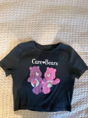 Svart ”Care Bears” croppad t-shirt - Cool svart/mörkgrå croppad t-shirt med Care Bears-tryck framtill i rosa och lila toner. T-shirten har rund hals och korta ärmar. Har klippt bort lappen tyvärr, men den är inköpt på Pull&Bear och passar mig som har strl S - ganska stretchig. Perfekt för dig som gillar retro och färgglada prints.