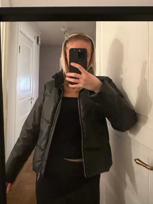 Svart pufferjacka i skinnimitation - Säljer en svart pufferjacka i skinnimitation med hög krage och dragkedja framtill. Jackan har en boxig passform och är quiltad för extra värme. Perfekt för dig som vill ha en stilren och trendig look under höst och vinter.