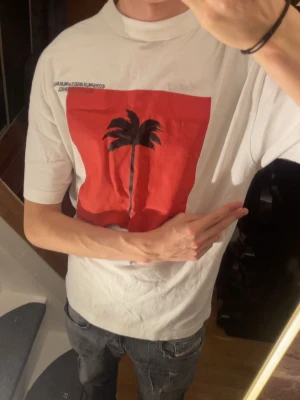 Palm Angels vit t-shirt - Palm angels tshirt ute‼️‼️ Skick 9/10✅✅Självklart äkta✅Kontakta📩 vid minsta fundering😌😌                                         Palm Angels t-shirt i vitt med stor röd fyrkant och svart palmtryck på framsidan. Tryck med text på både fram- och baksidan. Klassisk rund hals och korta ärmar. Materialet är mjuk bomull, perfekt för en chill streetwear-look.