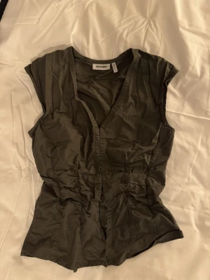 Olivgrön blus från Weekday - Säljer en olivgrön blus från Weekday i storlek 36. Blusen har v-ringning, korta ärmar och knappar framtill. Den är figursydd med snygga rynkade detaljer längs sidorna som ger en cool siluett. Perfekt att styla med jeans eller kjol.