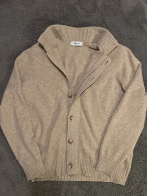 Gran Sasso ziptröja - Beige brun ziptröja från Gran Sasso med ribbade muddar och krage. Koftan har knappar framtill och är tillverkad i ett mjukt stickat material som passar perfekt för kyligare dagar. Enkel och stilren design som funkar till det mesta. Storleken är 52 vilket motsvarar ungefär M eller L. 