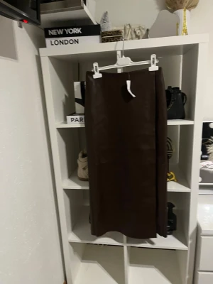 Brun lång skinnkjol med slits - Säljer en snygg brun lång kjol i skinnimitation med rak passform. Kjolen har en diskret slits framtill som ger extra edge och är fodrad med mjukt tyg på insidan. Perfekt för dig som gillar minimalistisk stil och vill ha något som sticker ut.