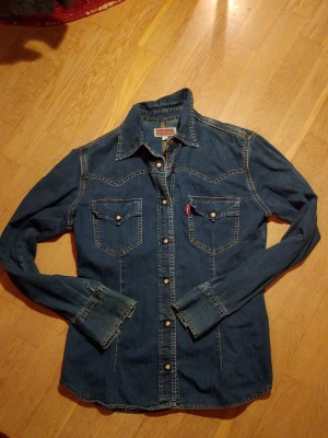 Levi's jeansskjorta med knappar - Snygg jeansskjorta från Camera jeansi mörkblå denim med klassiska kontrastsömmar. Skjortan har två bröstfickor med tryckknappar, lång ärm och markerad krage. Perfekt för dig som gillar en cool och avslappnad stil. Märket påminner mycket om levis med liknande färger.