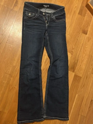 Mörkblå bootcut jeans med kontrastsömmar - Snygga mörkblå jeans med bootcut passform och tydliga vita kontrastsömmar. Framtill finns två fickor med lock och knappdetalj. Jeansen har klassisk femficksdesign och är tillverkade i jeansmaterial med en skön känsla.