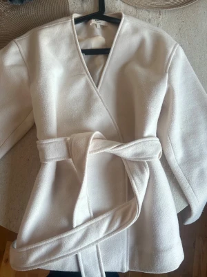Vit omlottjacka med bälte från Gina Tricot - Säljer en stilren vit omlottjacka från Gina Tricot med bälte i midjan. Jackan har en mjuk och lite strukturerad yta, långärmad och med en snygg v-ringad hals. Perfekt för dig som gillar minimalistisk och clean stil.