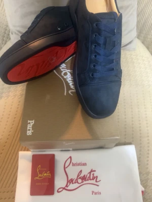Mörkblå Christian Louboutin sneakers - Snygga mörkblå sneakers från Christian Louboutin med klassisk röd sula och rund tå. Skorna har snörning och är tillverkade i mocka för en lyxig känsla. Perfekt för dig som vill sticka ut med exklusiva detaljer och ikonisk design.