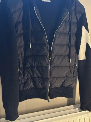 Svart pufferjacka Moncler med huva - Svart pufferjacka från Moncler med stickade ärmar och stor vit M-detalj på ena ärmen. Jackan har huva med snörning, dragkedja framtill och ribbade muddar. Materialmix av polyester och ull ger både värme och stil. Perfekt för dig som vill ha något unikt och snyggt.