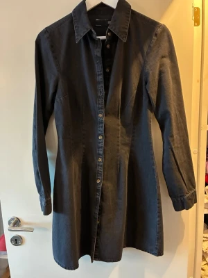 Svart jeans/skjortklänning från ASOS DESIGN - Säljer en svart jeans/ skjortklänning från ASOS DESIGN i 100% bomull. Klänningen har lång ärm, klassisk krage och knappar hela vägen framtill. Snygg, enkel och lätt att styla med olika accessoarer. Ger snygg timglasformad figur. Den är ca 86-88 cm lång. Aldrig använt då den är lite för tight för mig upptill. 