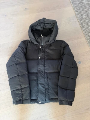 Svart Moncler pufferjacka med huva - Säljer en svart pufferjacka från Moncler med huva och stora fickor framtill. Jackan har dragkedja och tryckknappar, samt ribbade muddar på insidan. Klassisk Moncler-logga på ärmen och justerbar huva. Perfekt för kalla dagar.