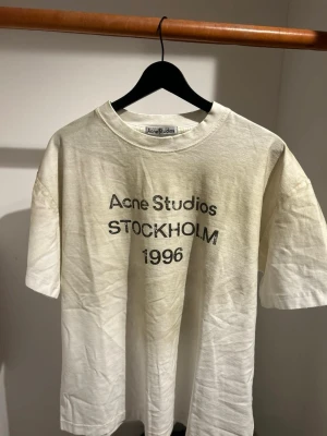 Vit Acne Studios t-shirt med tryck - Säljer en vit t-shirt från Acne Studios med svart tryck 'Acne Studios STOCKHOLM 1996' på framsidan. T-shirten har rund hals och korta ärmar, tillverkad i mjuk bomull. Perfekt för dig som gillar clean och stilren design med en touch av street.